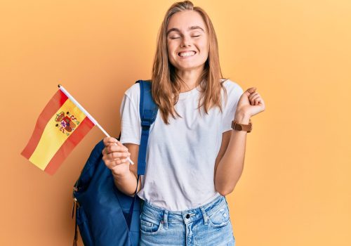 Beautiful blonde woman exchange student holding spanish flag screaming proud, celebrating victory and success very excited with raised arm