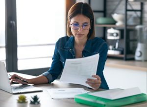 Pretty young business woman working with computer while consulting some invoices and documents in the kitchen at home