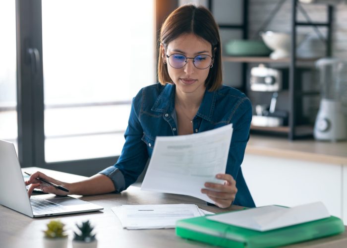 Pretty young business woman working with computer while consulting some invoices and documents in the kitchen at home